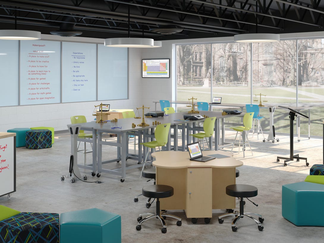 Modern STEM classroom with white presentation board, gray trapezoid tables arranged in clusters, teal soft seating cubes, wood-grain storage cabinet, and black ceiling with industrial lighting