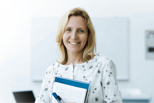 A smiling female educator holding a notebook, standing in front of a whiteboard