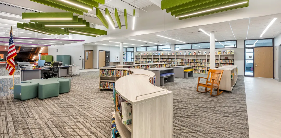 A modern, well-lit elementary school library with curved bookshelves, green acoustic ceiling panels, comfortable seating areas, and an American flag, showcasing a student-centered learning environment at Beatrice Elementary.
