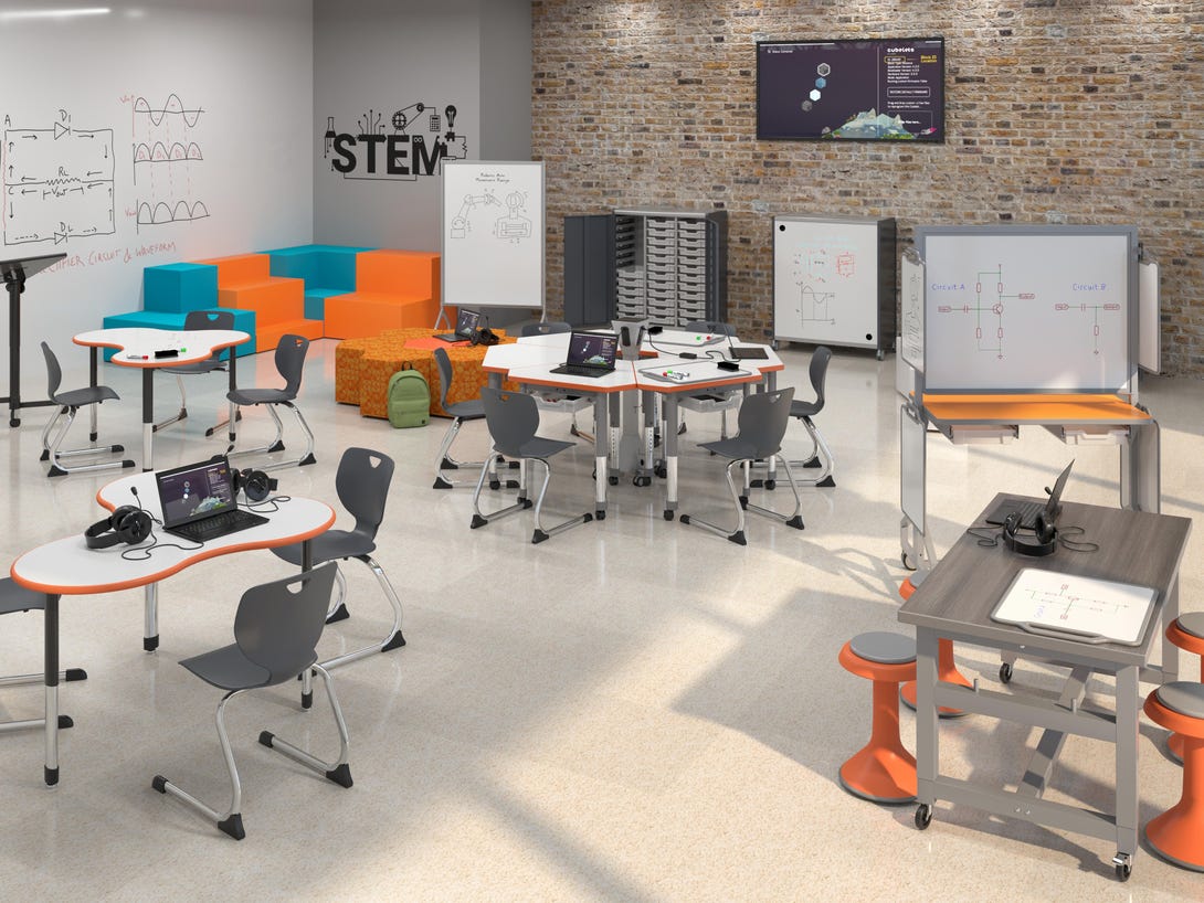 Industrial-style makerspace with exposed brick wall displaying 'STEM' signage, featuring angular orange and gray collaborative tables, mobile whiteboards, task chairs, and wall-mounted monitor